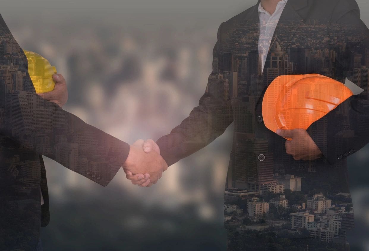 Two men shaking hands with a construction hat on their head.